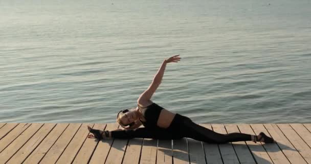 Žena dělá podélné rozdělení sedí na molu u řeky a naklání se k noze. — Stock video