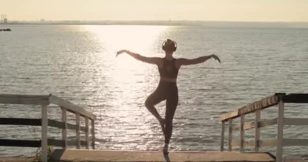 Mulher desportiva está fazendo ioga árvore pose de pé no cais perto do rio ao pôr do sol. — Vídeo de Stock