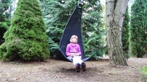 Маленькая красивая девочка на черном кресле-качалке висит на ветке дерева в лесистой усадьбе. . — стоковое видео