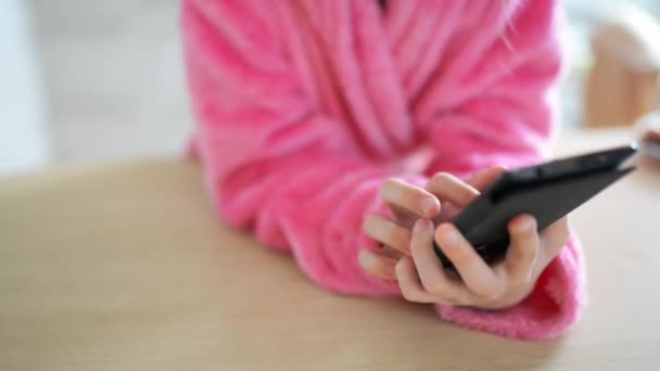 Маленькая красивая девушка в розовом халате и тряпичном бигуди сидит за столом на кухне и играет на своем мобильном телефоне . — стоковое видео