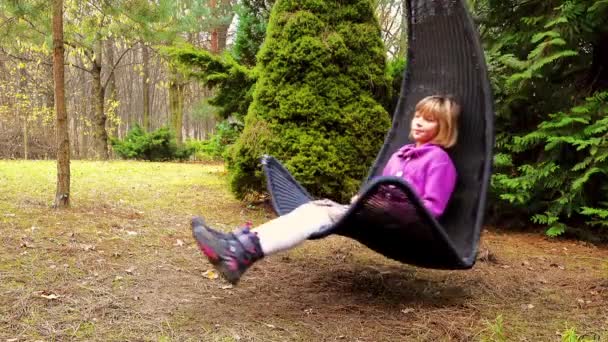 Маленькая красивая девочка на черном кресле-качалке висит на ветке дерева в лесистой усадьбе. . — стоковое видео