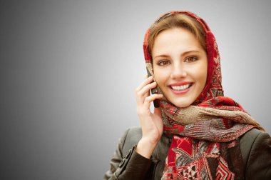 Genç güzel gülümseyen kadın cep telefonu gri arka plan üzerinde izole karışık kırmızı başörtüsü görüşmelerde.