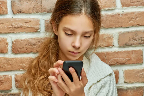 长头发的少女在砖墙的背景下在手机上打字 — 图库照片
