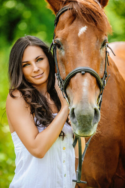 Woman in white dress keeps brown horse against background of summer green park.