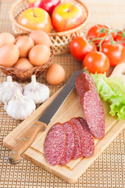 好吃的香肠和一把刀躺在切菜板上 柳条篮 柳条篮 鸡蛋和红色西红柿为背景 — 图库照片