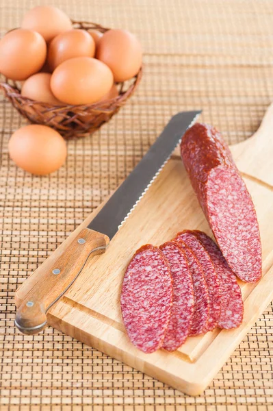 Tasty Sausage Knife Lying Cutting Board Wicker Basket Eggs Background — Stock Photo, Image