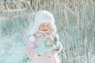 Küçük sevimli gülümseyen kız kış sihirli ormanda ayakta ve Noel ve yeni yıl dekorasyon top onun elinde tutan pembe ceket