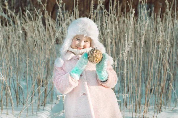 小可爱的微笑的女孩在粉红色的外套站在冬季神奇的森林 拿着圣诞节或新年装饰球在她的手中 — 图库照片