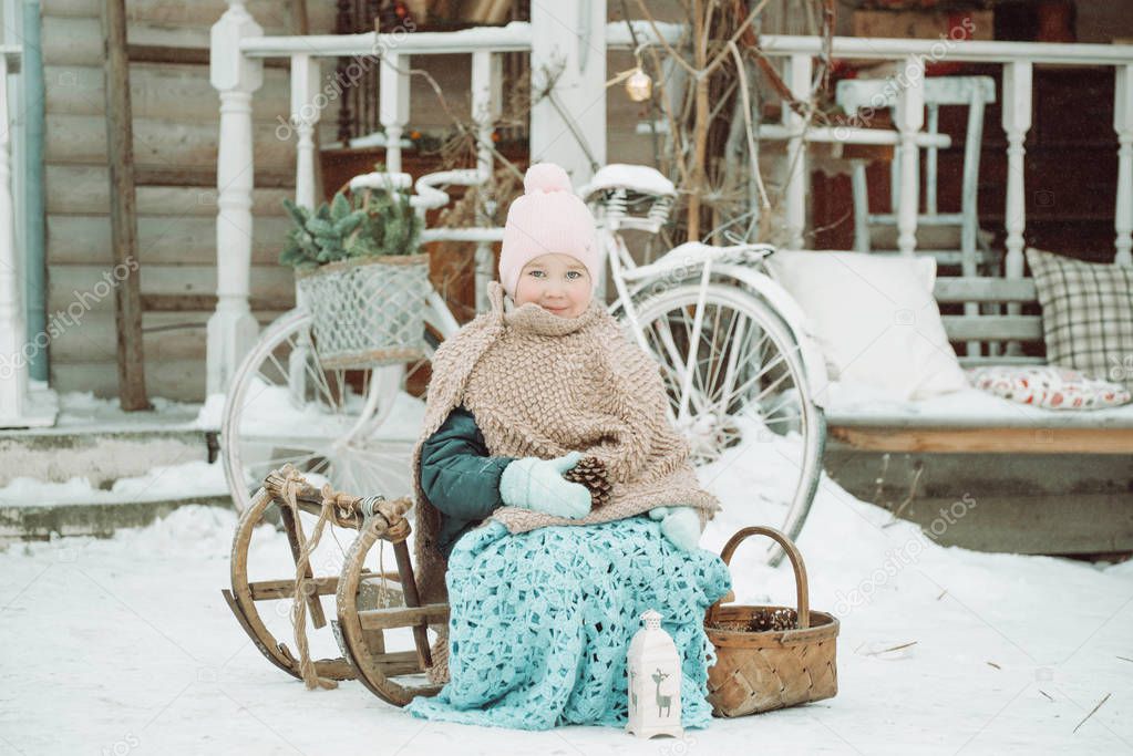 Little girl sitting on a sled in the winter, wrapped in a blanket, holding a lump, a festive atmosphere