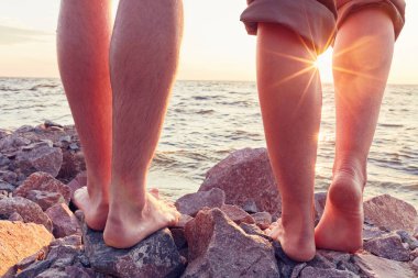 Bacakları sahilde. Ayak spası. Gün batımında sahilde sarılıp öpüşen genç bir çift. İki sevgili, erkek ve kadın, suyun yanında yalınayak. Yaz aşkı.