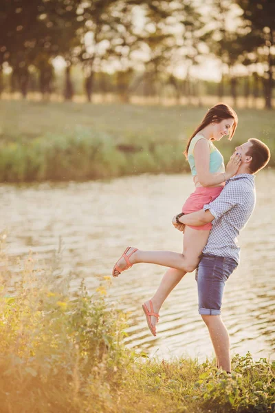 Verliebte in einem See. Junges verliebtes Paar sitzt auf dem Parkgelände am Wasser, während dieser junge Mann im Sonnenuntergang Gitarre spielt. — Stockfoto