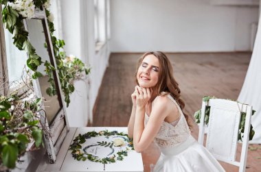 Beyaz elbiseli güzel bir gelin pencerenin yanındaki sandalyede oturuyor ve bir düğün buketi tutuyor..