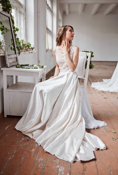 Uma noiva bonita em um vestido branco está sentado em uma cadeira perto da janela e segurando um buquê de casamento . — Fotografia de Stock