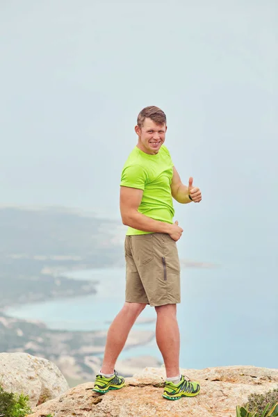 Turista em uma camiseta verde no topo da montanha mostra polegar para cima — Fotografia de Stock