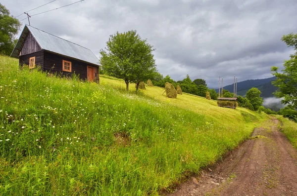 Panorama de montagne d'été. Petite maison en bois chalet et grange sur vallée verdoyante de montagne sur ciel brumeux lumineux, nuages et montagne — Photo