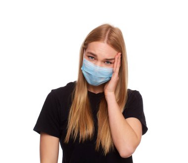 Kapalı gözlü, tıbbi grip maskesi takan, başını eliyle tutarak beyaz arka planda izole edilmiş genç sarışın kızın stüdyo portresi..