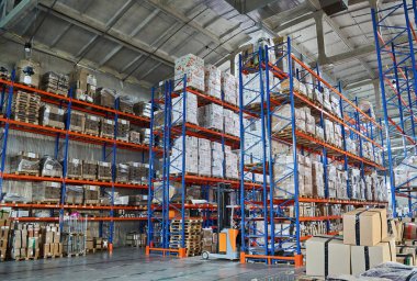 Spacious warehouse interior with very tall blue and orange racking systems heavily stocked with palletized goods and boxes. A forklift operates in an aisle.