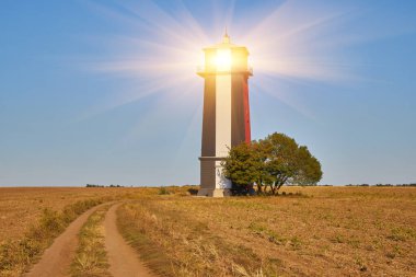 Uzun, çizgili bir deniz feneri (kırmızı, beyaz ve muhtemelen siyah renkli bantlar) açık, açık ve kuru bir alanda açık mavi bir gökyüzünün altında duruyor. Yanında tek bir ağaç var..