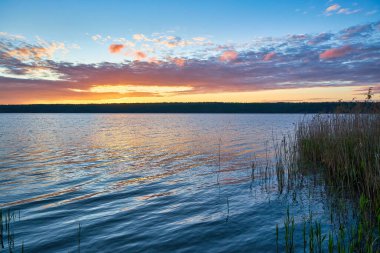 Muhteşem bir günbatımı, sakin bir göl üzerinde ateşli portakallar, kırmızılar ve sarılar ile gökyüzünü boyar. Ufukta görünen parlak güneş, suya parlak bir yansıma yayar..