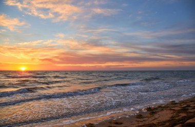Okyanus üzerinde gün doğumunun geniş açılı görüntüsü. Dalgalar yavaşça kumlu bir plaja çarpar ve gökyüzünde yumuşak pastel renkler oluşur..