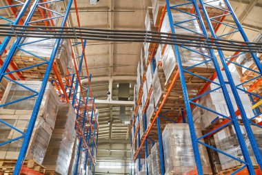 View looking up between towering blue and orange pallet racks in a warehouse. The high shelving is heavily stocked with shrink-wrapped goods. High-density industrial storage.