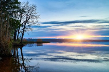 Sakin bir göl, akşam gökyüzünün yumuşak pastel renklerini mükemmel bir şekilde yansıtır. Günbatımında pembe, portakal ve sarılarla boyanır. Siluetli sazlıklar ve uzak bir ağaç hattı huzur dolu sahneyi tamamlıyor..