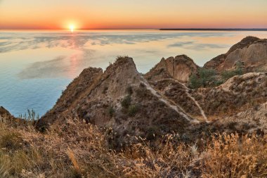 Önünde engebeli uçurumlar ve kuru otlar olan bir kıyı şeridi ve ufka uzanan sakin bir deniz ya da okyanus manzarası..