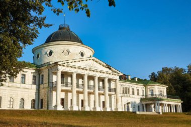 The grand neoclassical facade of Kachanivka Palace in Ukraine, featuring a central dome and colonnaded portico, viewed across a wide, sun-dappled lawn.