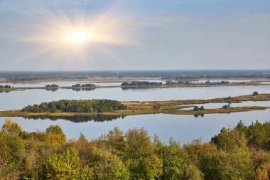 Geniş bir nehir manzarası. Açık bir gökyüzünün altında ağaçlar ve bitkilerle kaplı birden fazla ada var..