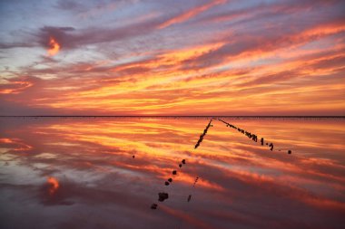 Sakin bir tuz gölünün üzerinde nefes kesici bir gün batımı. Kırmızı, turuncu ve pembe bulutlu canlı, ateşli gökyüzü durgun suda mükemmel bir şekilde yansıyor. Eski tahta direkler sıraya dizilir.