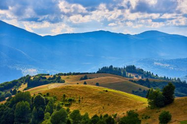 Bulutlu bir gökyüzünün altında dağ eteklerinin, köy evlerinin, ormanların ve yama alanlarının yer aldığı manzaralı yaz manzarası. Kırsal Karpatya görüşü.
