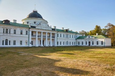 The grand neoclassical facade of Kachanivka Palace in Ukraine, featuring a central dome and colonnaded portico, viewed across a wide, sun-dappled lawn.