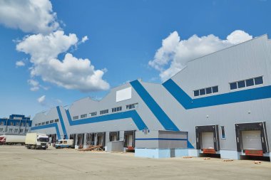 Exterior facade of a large, modern warehouse or distribution center with grey and blue metal siding, multiple loading docks, and roll-up doors, under a blue sky with white clouds. Industrial logistics facility.