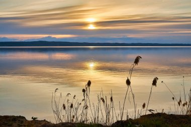 Sakin bir gölün üzerinde nefes kesen bir gün batımı, parlak güneş ufukta belirdiğinde suyun üzerinde parıldayan altın bir ışık yolu oluşturuyor. Huzurlu ve huzurlu.