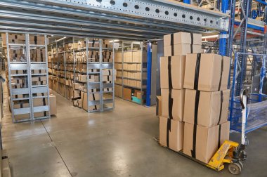 A yellow pallet jack loaded with a tall stack of sealed cardboard boxes stands in a warehouse aisle, surrounded by high metal shelving units packed with inventory.