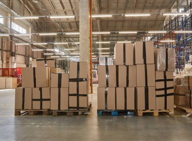 Interior of a large warehouse showing tall stacks of sealed cardboard boxes on wooden pallets, ready for shipping or storage. Industrial logistics and inventory.
