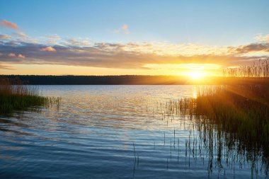Muhteşem bir günbatımı, sakin bir göl üzerinde ateşli portakallar, kırmızılar ve sarılar ile gökyüzünü boyar. Ufukta görünen parlak güneş, suya parlak bir yansıma yayar..