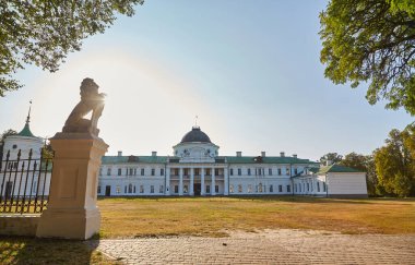View of Kachanivka Palace in Ukraine, with a silhouetted lion statue on a pedestal guarding the entrance. The historic manor is seen across a lawn under a bright sky.