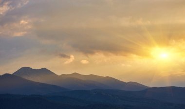Gün batımında veya gündoğumunda turuncu ve sarı parlayan bulutlarla dolu dramatik, desenli bir gökyüzüne karşı dağ tepeleri ve tepelerin yakın çekim silueti. Atmosferik.