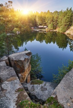Korostyshiv taş ocağı gölü manzaralı, kayalık önplanlı, ağaçları ve gökyüzünü yansıtan, ve su boyunca görünen yapılar..