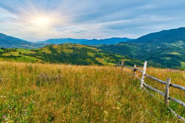 Kırsal ahşap çitler, dramatik bulutlarla dolu mavi gökyüzünün altındaki ormanlı tepelere bakan çimenli bir çayırlık yamaçtan geçiyor. Kırsal yaz manzarası.