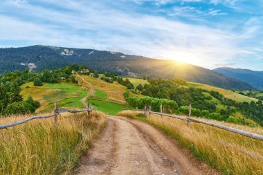 Yüksek, çimenli bir dağ yamacındaki ahşap bir çitin yanında uzanan toprak yolun manzarası. Açık mavi gökyüzünün altında görünen katmanlı orman tepeleri. Yürüyüş yolu.