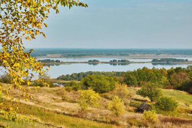 Kuru çimenler ve küçük ahşap bir kulübeyle sonbahar yamacından görünen adalarla geniş nehir manzarası. Huzurlu kırsal görünüm.