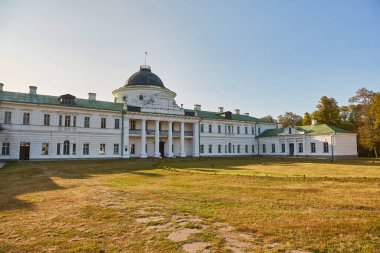 The grand neoclassical facade of Kachanivka Palace in Ukraine, featuring a central dome and colonnaded portico, viewed across a wide, sun-dappled lawn.