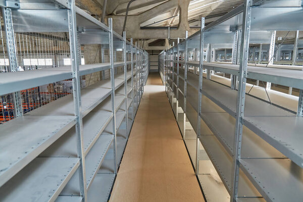 View down a long aisle in a warehouse or storage facility, lined with tall, empty metal shelving units. Industrial interior with high ceiling and concrete floor.