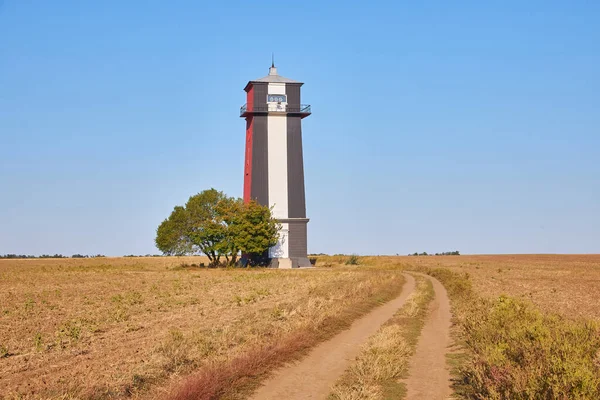 Uzun, çizgili bir deniz feneri (kırmızı, beyaz ve muhtemelen siyah renkli bantlar) açık, açık ve kuru bir alanda açık mavi bir gökyüzünün altında duruyor. Yanında tek bir ağaç var..