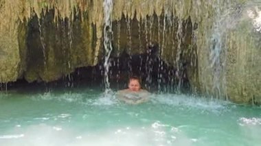 Mavi suyla dolu bir gölde şelalenin altında yüzen kızıl saçlı güzel bir kadın. 4K görüntü klibi