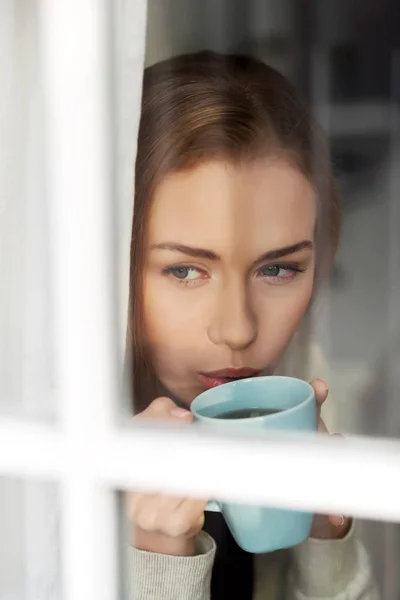 Krásná Kavkazský žena pít horkou kávu nebo čaj a dívat se skrz okno. — Stock fotografie