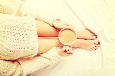 Genç bir kadın bir fincan kahveyle yatakta oturuyor.