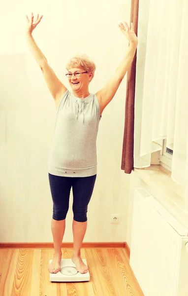 Gelukkig senior vrouw stond op gewicht schaal in woonkamer — Stockfoto